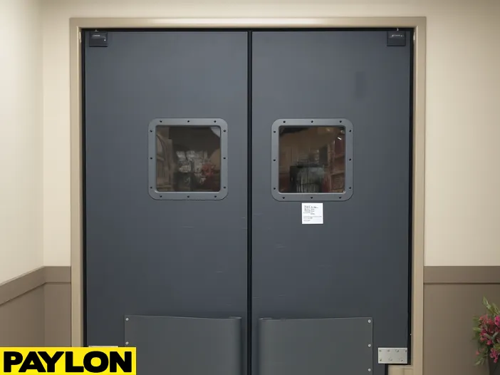 A closed PE-8000 industrial swinging door in a warehouse hallway A closed PE-8000 industrial swinging door in a warehouse hallway, designed for high-impact use with reinforced bumpers and square vision panels