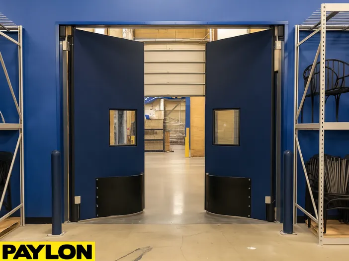Open PE-9000 industrial swinging doors in a warehouse setting Open PE-9000 industrial swinging doors in a warehouse setting, revealing a spacious storage area beyond. Designed for high-traffic zones and impact resistance