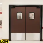 PE-720 retail swinging door installed in a school cafeteria hallway, featuring dark brown panels, stainless steel kick plates, and clear vision windows framed in black for safe two-way traffic flow