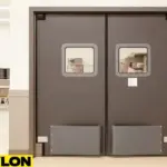PE-8000 industrial swinging door installed between a school cafeteria and kitchen, featuring dark impact-resistant panels, clear square vision windows, and lower bumpers for cart traffic in high-use areas
