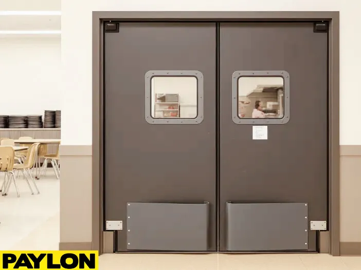 PE-8000 industrial swinging door installed between a school cafeteria and kitchen, featuring dark impact-resistant panels PE-8000 industrial swinging door installed between a school cafeteria and kitchen, featuring dark impact-resistant panels, clear square vision windows, and lower bumpers for cart traffic in high-use areas
