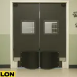Industrial Door PE-8000 double-swing doors installed in a veterinary clinic exam room, featuring square vision panels, kick plates, and a clean clinical setup with animal cages, paw print decals, and a stainless steel exam counter