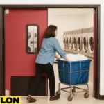 A woman pushes a blue laundry cart through the Industrial Door PE-8000 in a commercial laundromat, with red swinging doors providing quick access to the backroom and stainless steel washers visible in the background, perfect for efficient workflow in high-traffic utility areas
