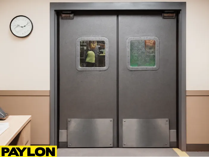Industrial Door PE-8000 swinging doors in a waste management facility, showing the transition from an office desk area to the warehouse floor Industrial Door | PE-8000 swinging doors in a waste management facility, showing the transition from an office desk area to the warehouse floor with workers and dumpsters visible through the vision panels