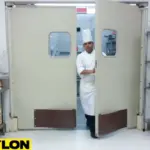 A chef in uniform looks through the opening of an off-white double swinging door in a restaurant kitchen