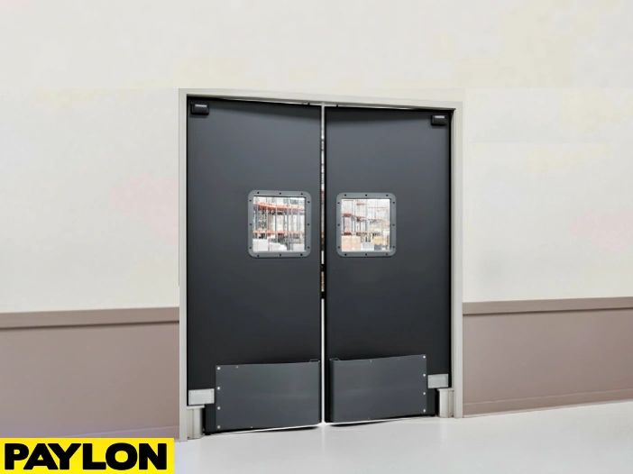 Heavy-duty black commercial swinging doors by Paylon installed in a warehouse facility, featuring kick plates and vision panels for safe, high-traffic access between storage and work areas Heavy-duty black commercial swinging doors by Paylon installed in a warehouse facility, featuring kick plates and vision panels for safe, high-traffic access between storage and work areas.