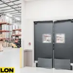 Heavy-duty industrial swinging doors by Paylon in a warehouse environment, featuring gray impact panels and windows for forklift and personnel access