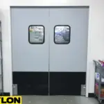 Light grey double-swing warehouse impact door with black reinforced panels in a storage area Light grey double-swing warehouse impact door with black reinforced panels in a storage area