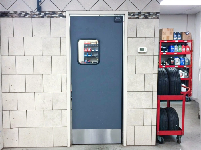 Single gray impact door with small window inside an auto repair shop, surrounded by tiled walls and shelving with part Single gray impact door with small window inside an auto repair shop, surrounded by tiled walls and shelving with parts