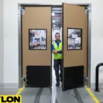 Worker passing through tan double-swing warehouse impact door with reinforced lower panels Worker passing through tan double-swing warehouse impact door with reinforced lower panels