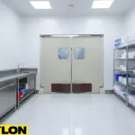 Beige double clean room doors with kick plates, shelves of supplies, and hanging lab coats in a sterile laboratory space.