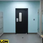 Black single swinging door in a clean locker room hallway with gray lockers and shower stalls.