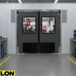 Gray factory swinging door with vision panels installed between two high-traffic workstations in an assembly line environment