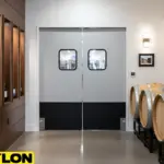 Light gray swinging doors with dual vision windows installed between a winery production area and tasting hallway.
