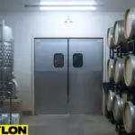 Stainless steel swinging doors in a winery cellar with wine barrels and fermentation tanks on both sides.